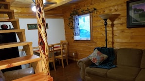 Ein gemütliches Blockhaushaus-Interieur mit einer Holztreppe, einer Couch, einem Esstisch mit Stühlen und einem Fenster mit Vorhängen.