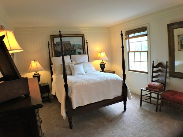 A four-poster bed with white linens, two bedside lamps, a chair with a red cushion, and a painting above the bed. - The Manor House Country Home and Retreat (East Peoria)