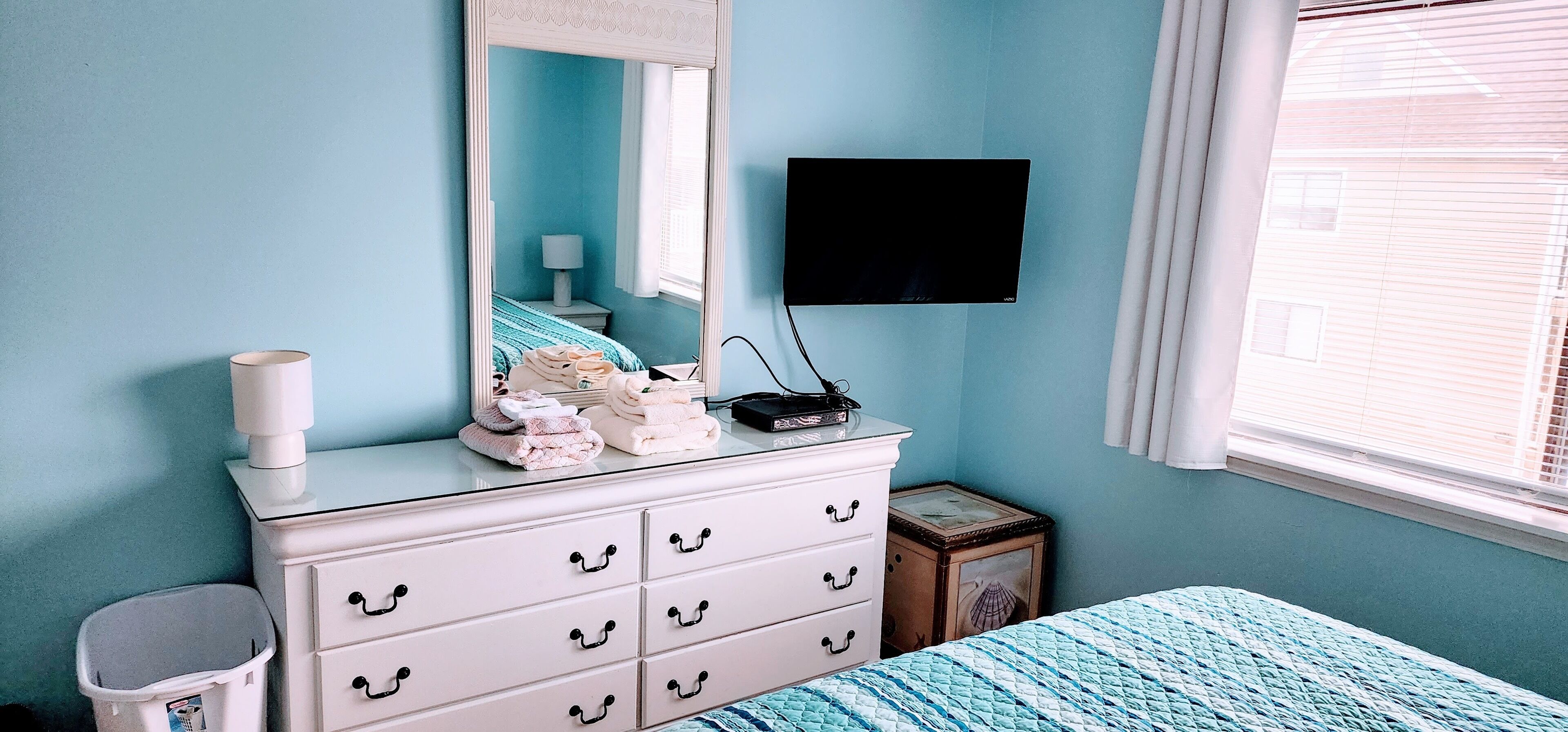 Une chambre à coucher comprenant un lit, une commode avec des tiroirs, un miroir, un téléviseur et une fenêtre avec des stores.
