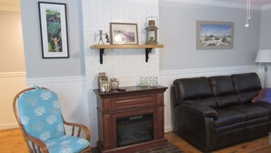 Een woonkamer met een blauwe gestreepte fauteuil, een donker leren bank, een open haard en decoratieve objecten op een plank.