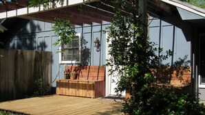 Een houten terras met een bankje, een potplant en een raam met zonwering.