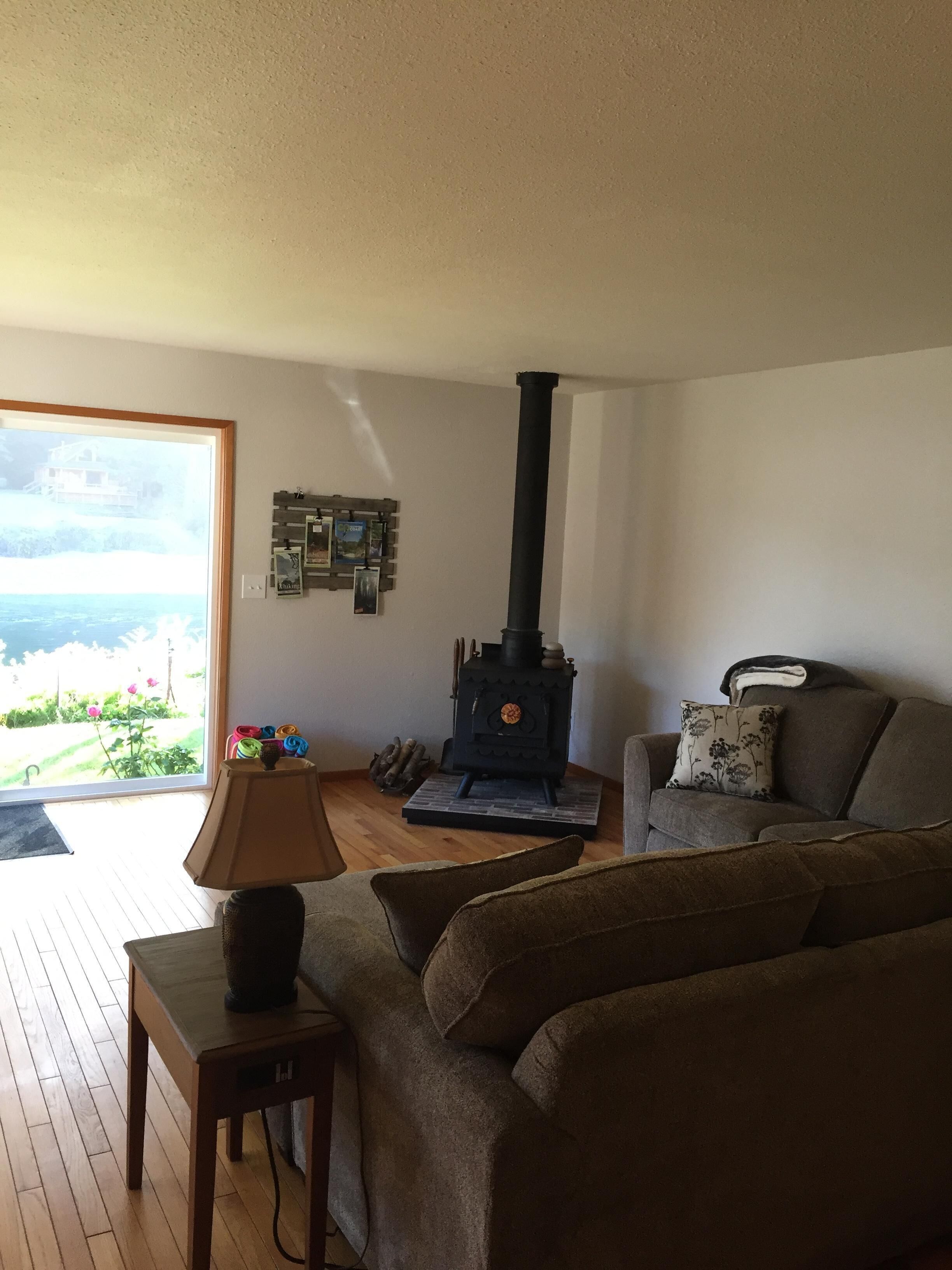 Una sala de estar con un sofá, una mesa con una lámpara, piso de madera, estufa negra y vista a través de un ventanal.