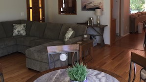Una sala de estar con un sofá modular, una mesa redonda con una planta en maceta, un candelabro colgante y vista a la cocina.