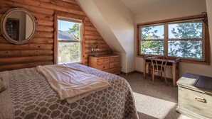Ein Schlafzimmer mit Bett, Schreibtisch, Stuhl, Kommode und einem großen Fenster mit Blick auf Bäume und Berge.