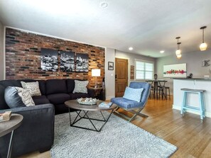 A living room with a brick wall, a sofa, a coffee table, and a chair. There is a dining area with a table and chairs in the background.