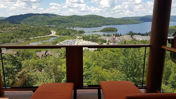 A balcony with a view of a lake, surrounding greenery, and distant buildings. - Tremblant Prestige - Equinoxe 140-1 (Mont-Tremblant)