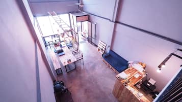 A spacious loft-style room with a high ceiling, a dining area with a long table and chairs, a seating area with a bench, and a kitchenette with a wooden counter.