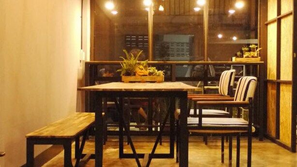 A cozy dining area with a wooden table and benches, a potted plant on the table, and hanging lights. - Asleep Hostel (Kanchanaburi)