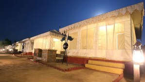 A row of traditional tents with a central signpost, a small table, and steps leading to the entrance. - Chokhi Dhani Desert Camp (Jaisalmer)