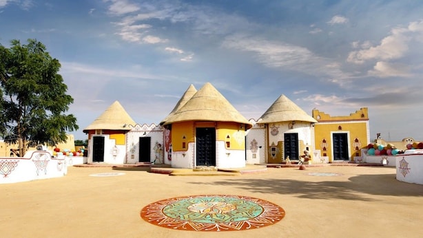 A courtyard with three traditional conical-roofed buildings, a large decorative circle in the center, and a tree to the left. - Chokhi Dhani Desert Camp (Jaisalmer)