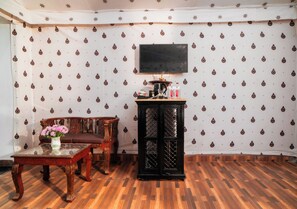 Una habitación con papel tapiz estampado, piso de madera, televisor montado en la pared, un mueble de madera y una mesa con un florero.
