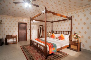 A four-poster bed with a colorful bedspread, a wooden nightstand with a vase, and a patterned wall.