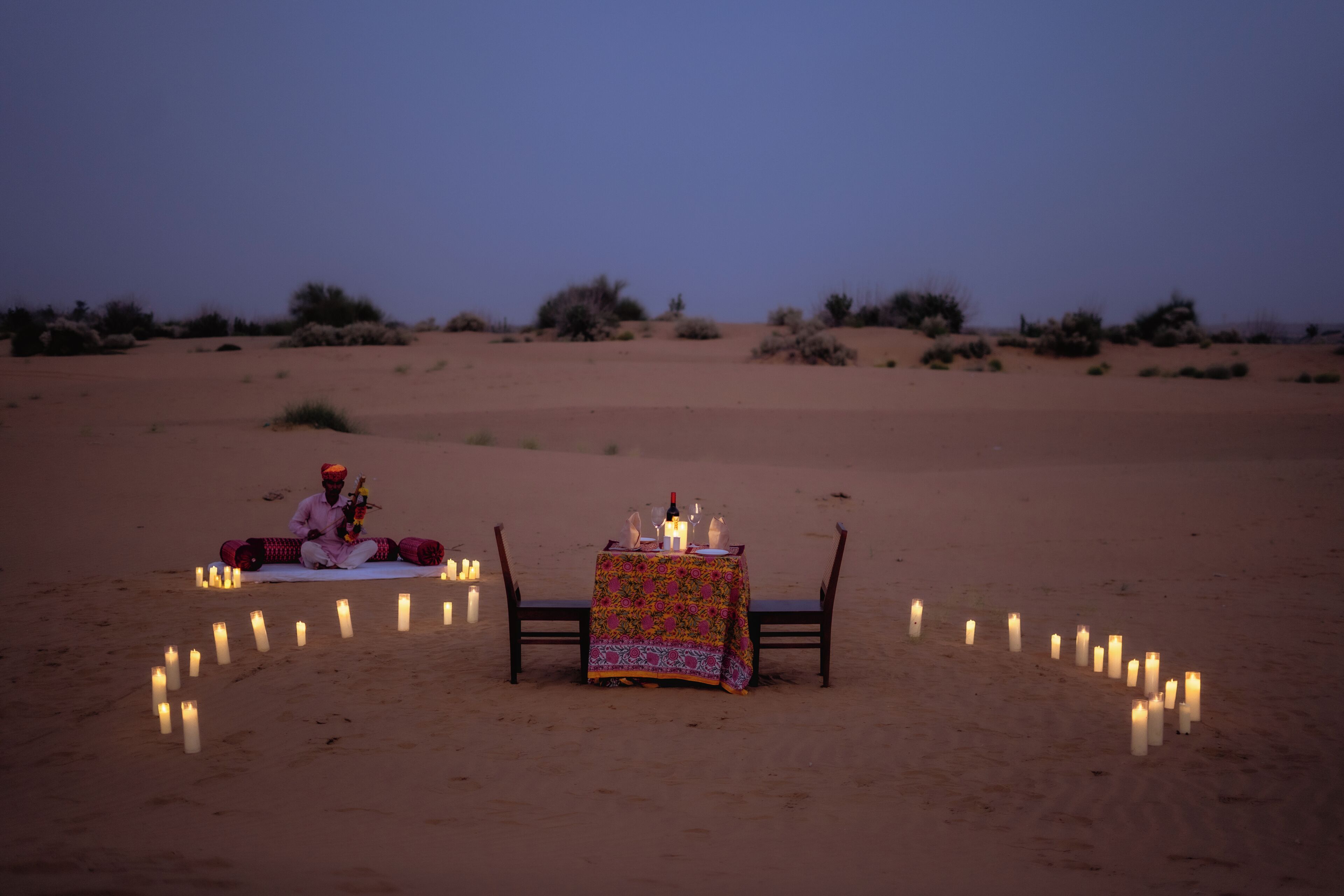 A desert scene with a table set for two, surrounded by candles, and a person sitting on the sand.