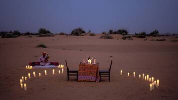 A desert scene with a table set for two, surrounded by candles, and a person sitting on the sand.