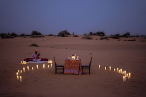 A desert scene with a table set for two, surrounded by candles, and a person sitting on the sand.