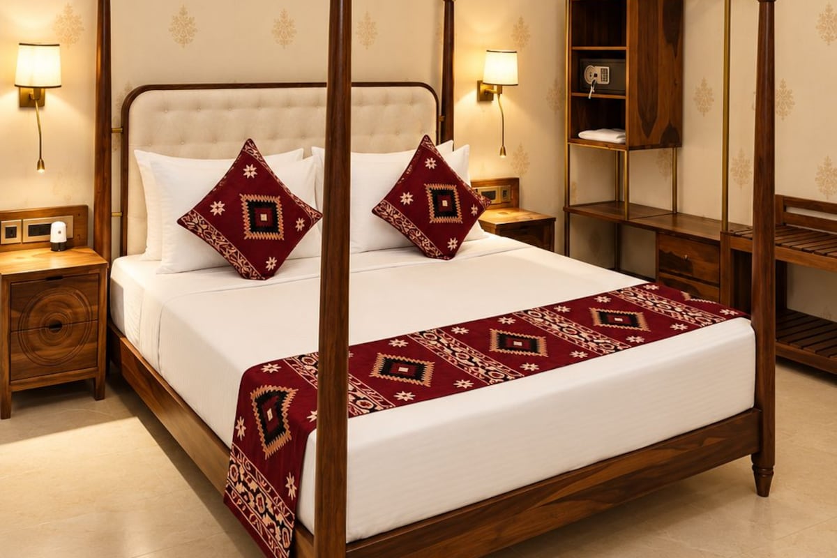 A four-poster bed with a wooden frame, two matching pillows, and a red patterned bedspread. There are two bedside tables with lamps, a wooden nightstand, and a wooden wardrobe with a mirror.