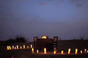 A table set for two with candles and a bottle of wine, surrounded by sand dunes at dusk.