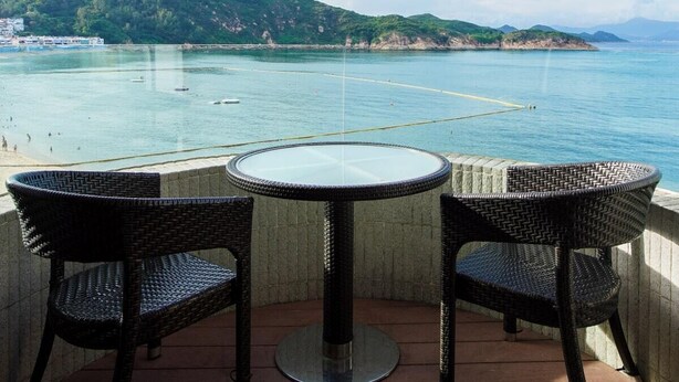 A balcony with a round table and two wicker chairs, overlooking a coastal area with boats and a hilly landscape. - The Warwick (Cheung Chau)