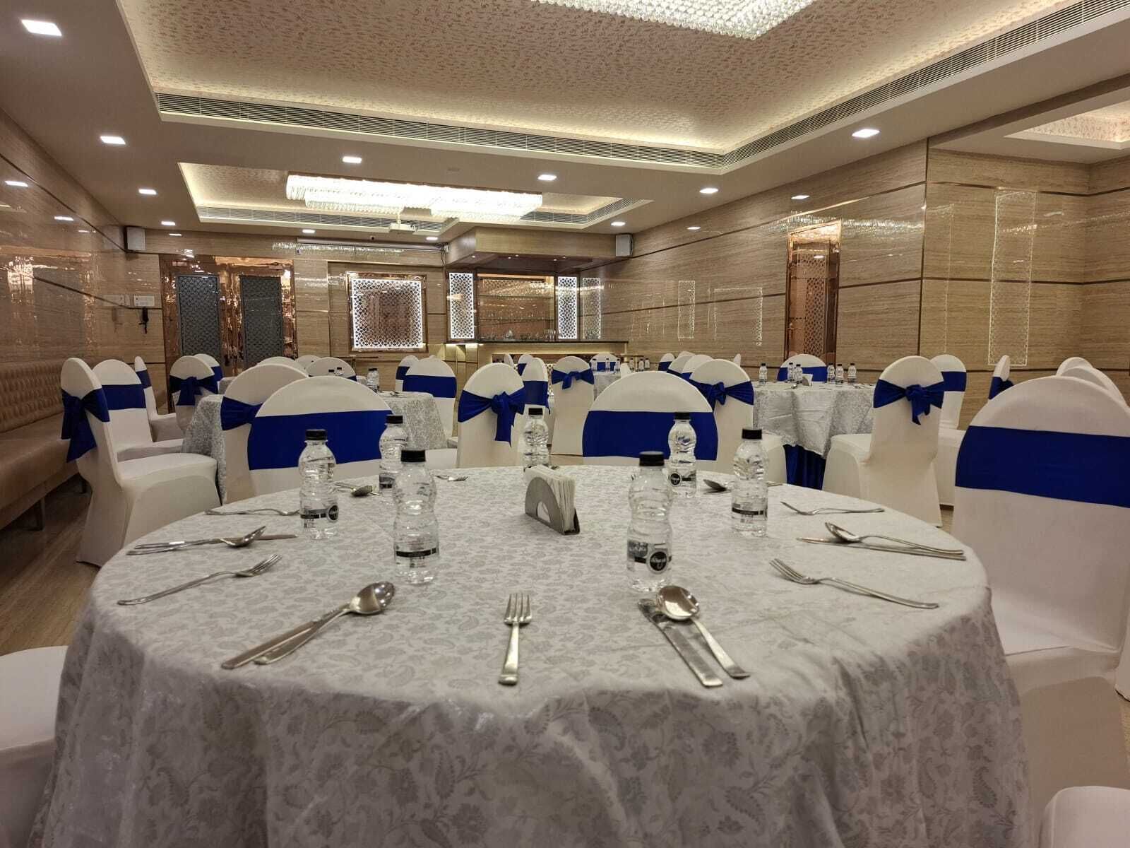 Un salón de banquetes con mesas redondas, sillas blancas adornadas con cintas azules y mantel de estampado de mármol.