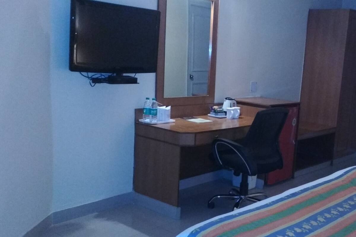 Habitación de hotel con un escritorio de madera, una silla de oficina negra, una cama con manta a rayas, un televisor montado en la pared y un armario.