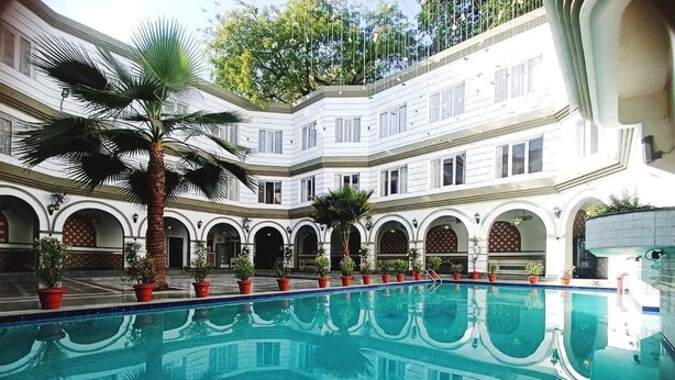 A pool with clear blue water, surrounded by potted plants and palm trees, with a building featuring arched windows and balconies in the background. - Grand Continental Hotel  (Prayagraj)