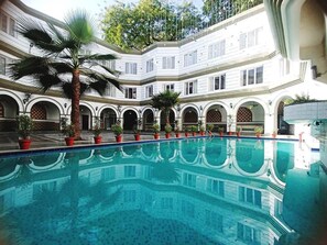 Una piscina con agua azul cristalina, rodeada de plantas en macetas y palmeras, con un edificio de fondo que presenta ventanas arqueadas y balcones.