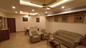 A living room with a sofa, chairs, a dining table, and ceiling fans. - Grand Continental Hotel  (Prayagraj)
