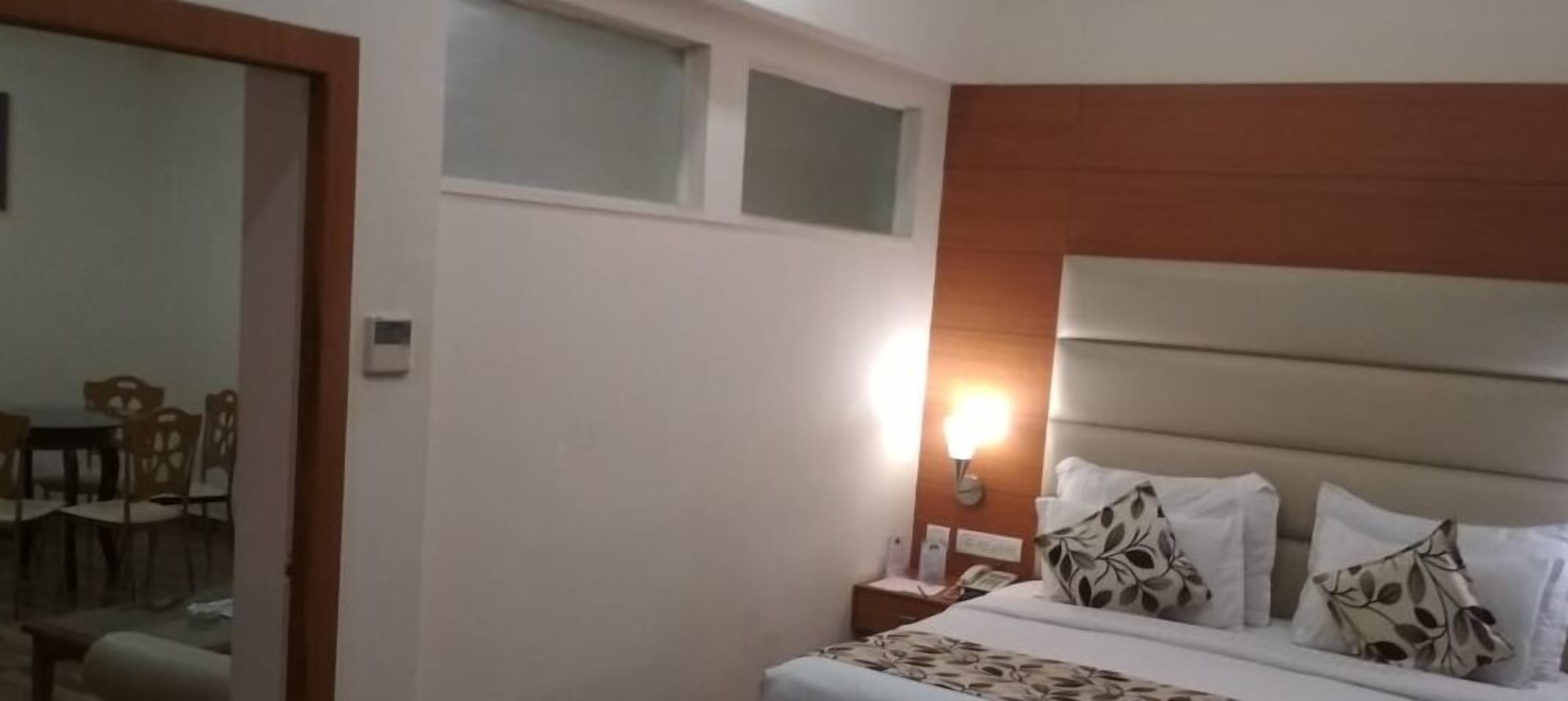 A hotel room with a bed, a nightstand, a wall-mounted lamp, and a dining area visible through a door.