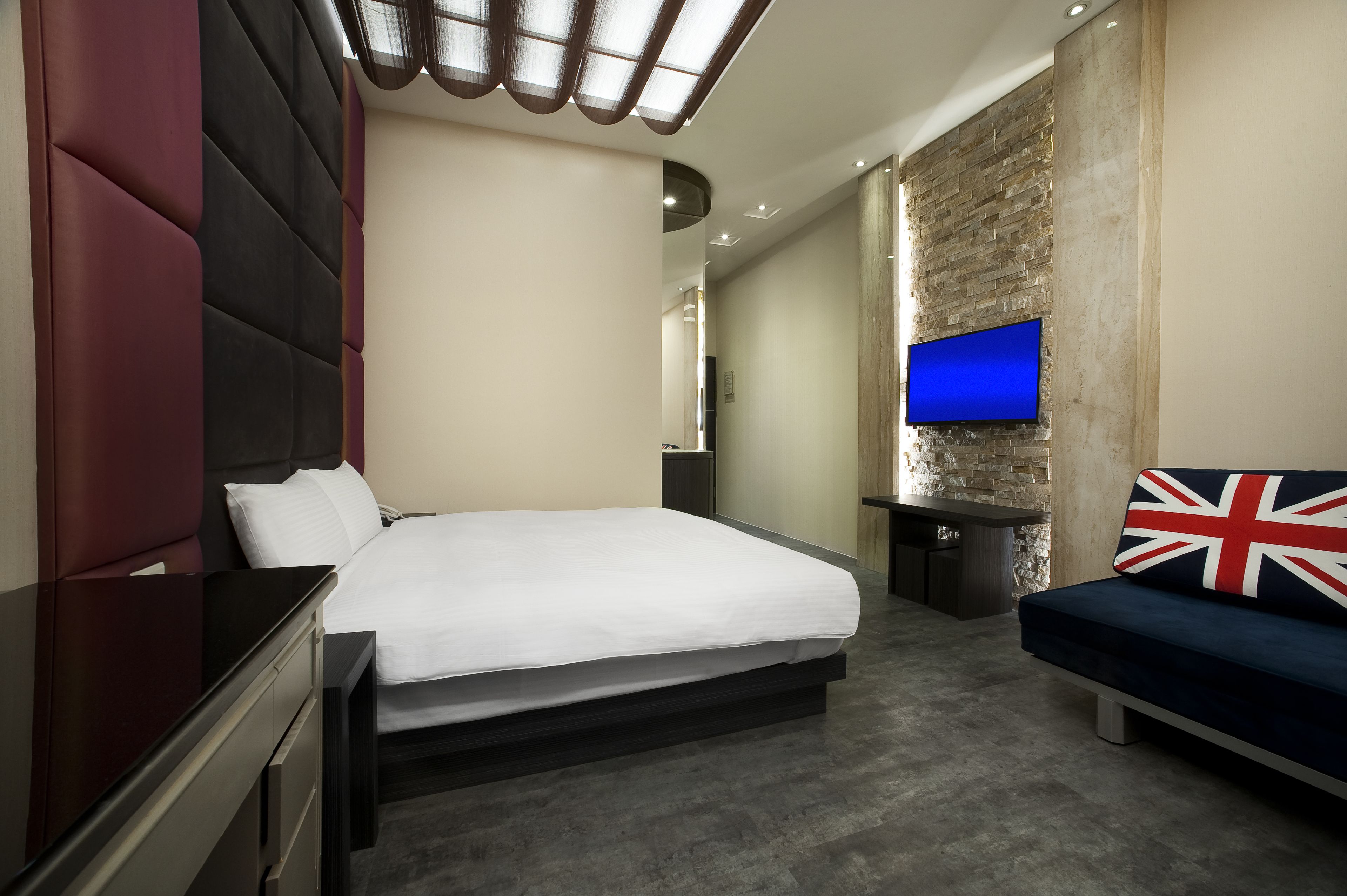 Une chambre d’hôtel moderne avec un grand lit, un téléviseur fixé au mur en briques, un canapé agrémenté d’un coussin orné de l’Union Jack et un petit bureau.