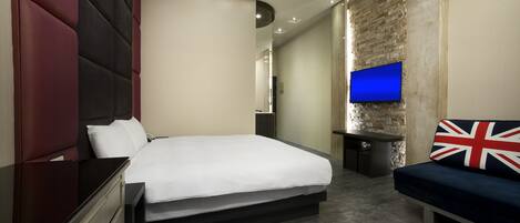 Une chambre d’hôtel moderne avec un grand lit, un téléviseur fixé au mur en briques, un canapé agrémenté d’un coussin orné de l’Union Jack et un petit bureau.
