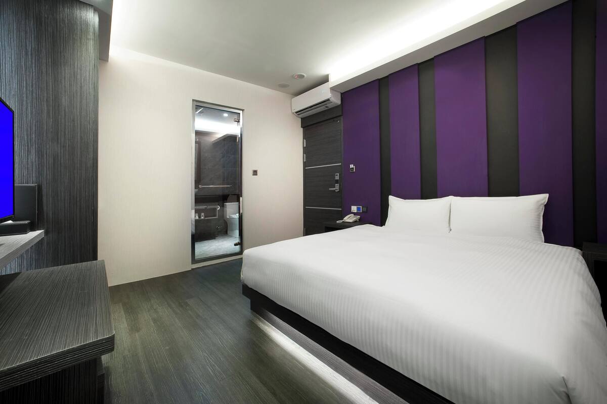 A modern hotel room with a large bed, a desk, and a wardrobe.