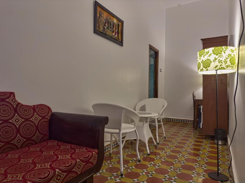 Una habitación con un sofá estampado, sillas blancas, una lámpara de pie y un suelo de baldosas.