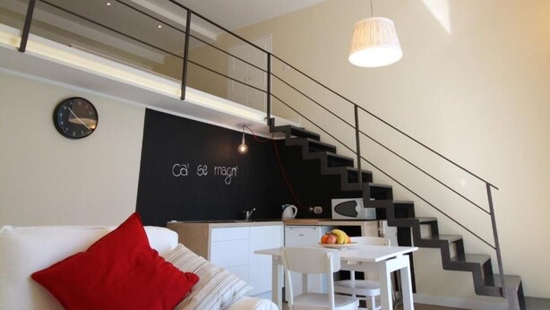 A modern living space with a staircase, a white sofa with red cushions, a dining table, and pendant lights.