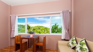 A room with a wooden floor, a table with chairs, a sofa with cushions, a window with curtains, and a view of greenery outside. - Greenery Resort Koh Tao (Koh Tao)