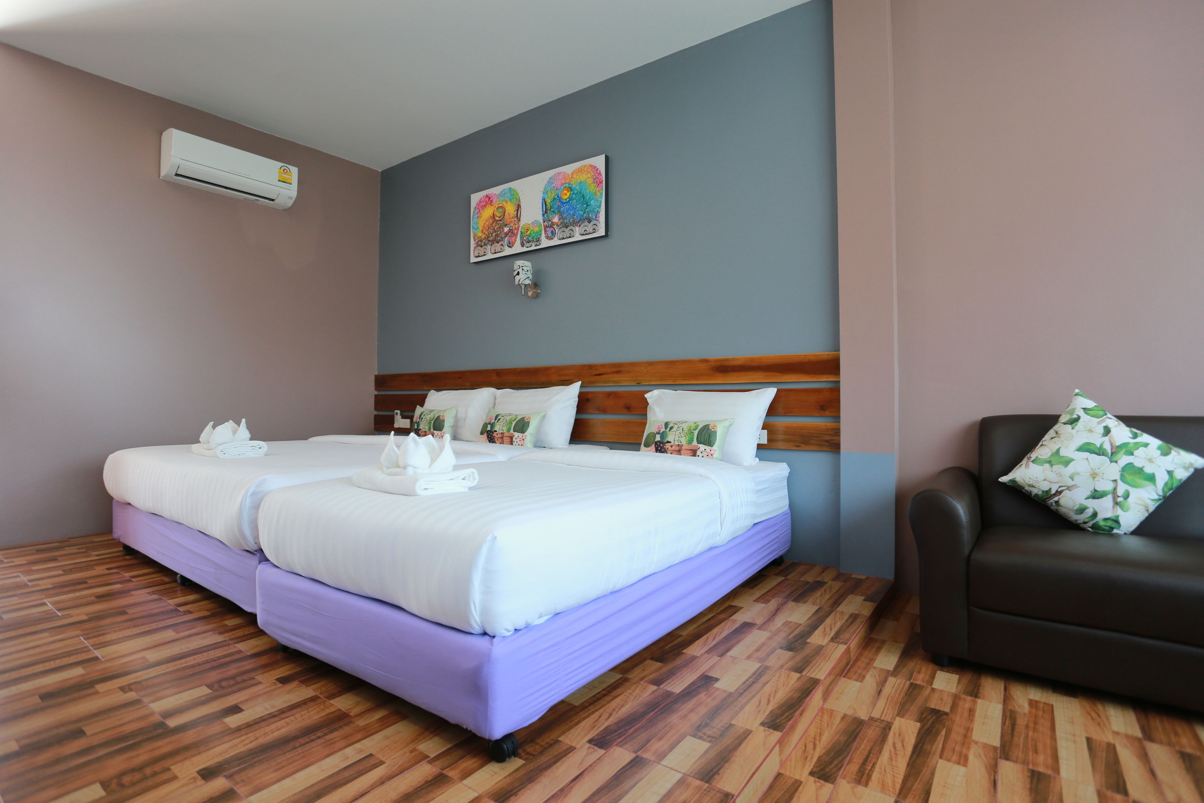 A hotel room with a double bed, a wooden headboard, a brown leather sofa, and a wall-mounted air conditioner.