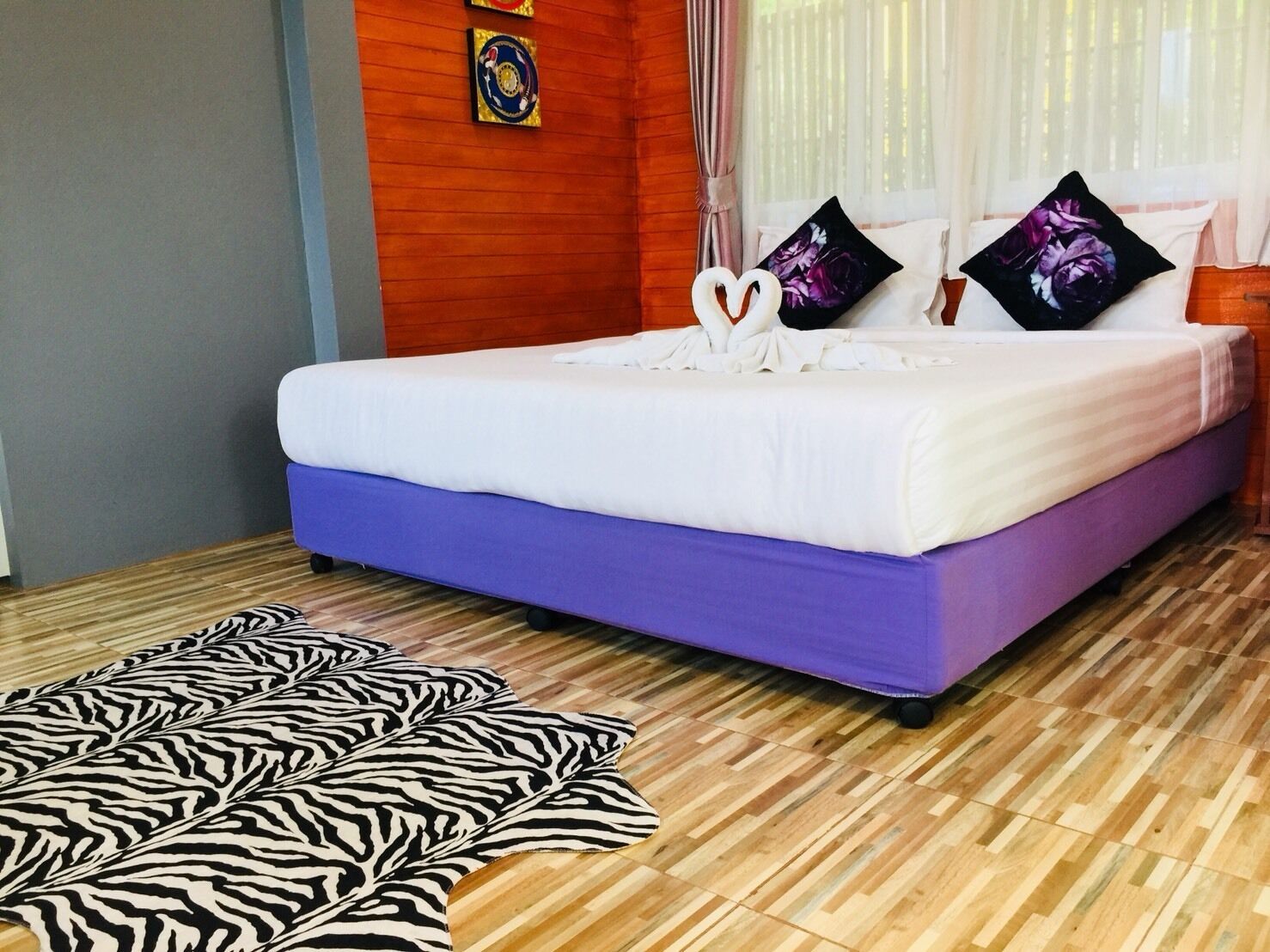 Ein Bett mit violetter Basis und weißer Bettwäsche, zwei Zierkissen, ein Teppich mit Zebra-Muster und ein Holzboden.