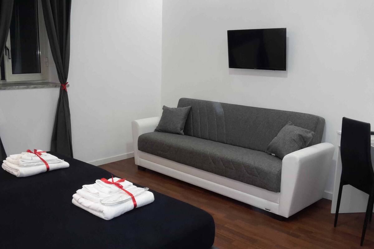 Una habitación de hotel moderna con un sofá gris, un edredón negro y un televisor de pantalla plana montado en la pared.