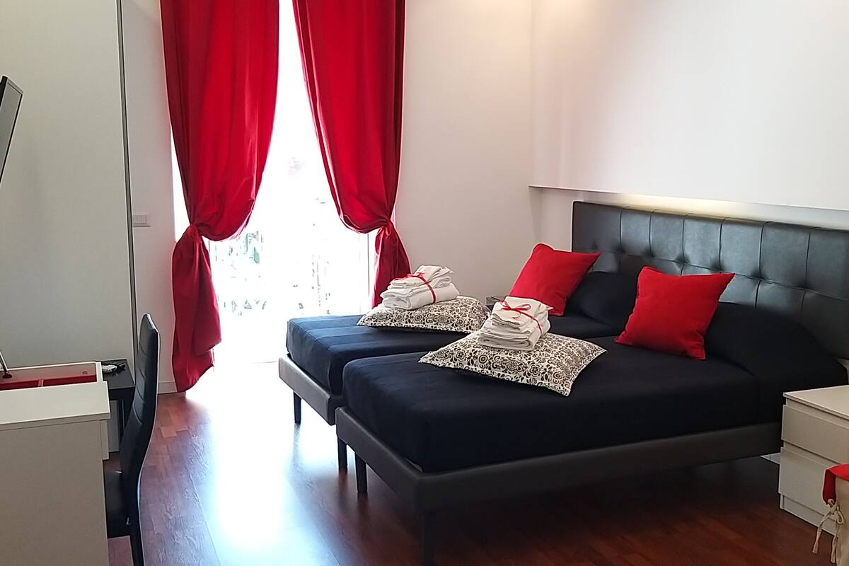 Un dormitorio con dos camas, cortinas rojas, una mesita de noche y un televisor montado en la pared.