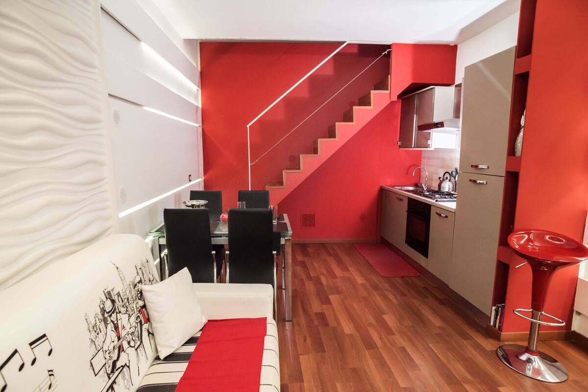 Una sala de estar moderna con sofá, mesa de comedor y detalles en rojo. Se ve una cocina con pared roja y una escalera al fondo.