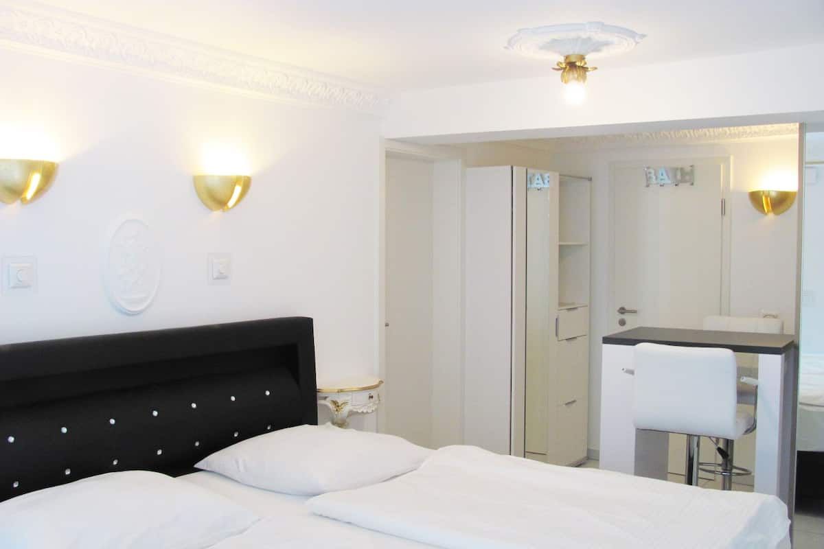 Ein Hotelzimmer mit einem Bett, einem Schreibtisch und einem Stuhl. Ein Spiegel und eine Tür zu einem weiteren Raum sind vorhanden.