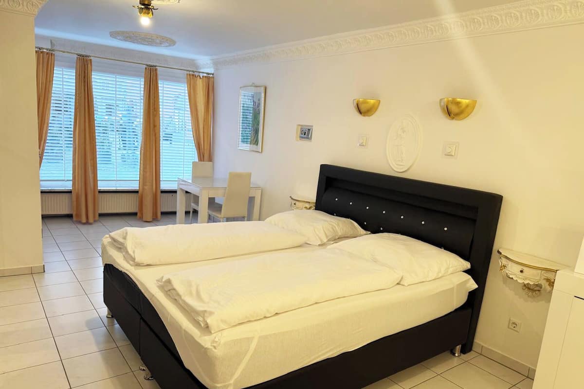 Ein Doppelbett mit weißen Leinentüchern, ein kleiner Schreibtisch mit Stuhl und ein Fenster mit Vorhängen.