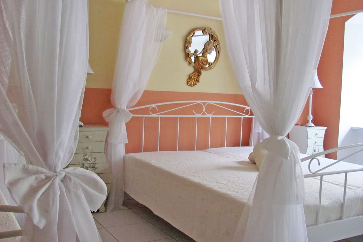 Ein Schlafzimmer mit einem Bett, weißen Vorhängen, einem Nachttisch, einer Kommode und einem Spiegel an der Wand.
