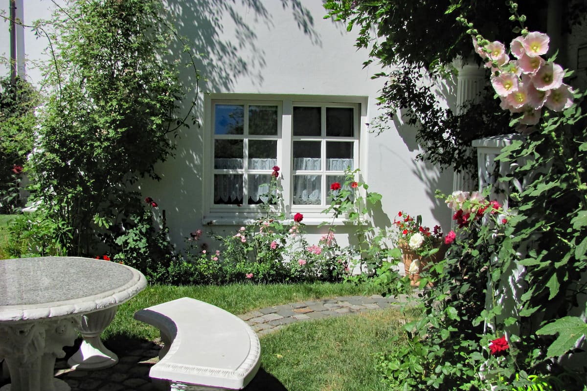 Ein Garten mit einem Steintisch und einer Bank, ein weißes Gebäude mit einem Fenster und verschiedenen Blumen.