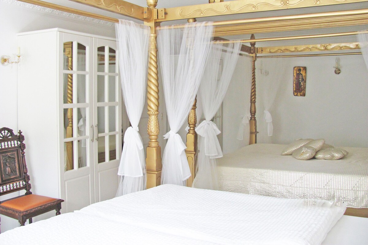 Una cama con dosel, con cubrecamas blancos y dosel, una silla de madera y una puerta blanca con paneles de vidrio.