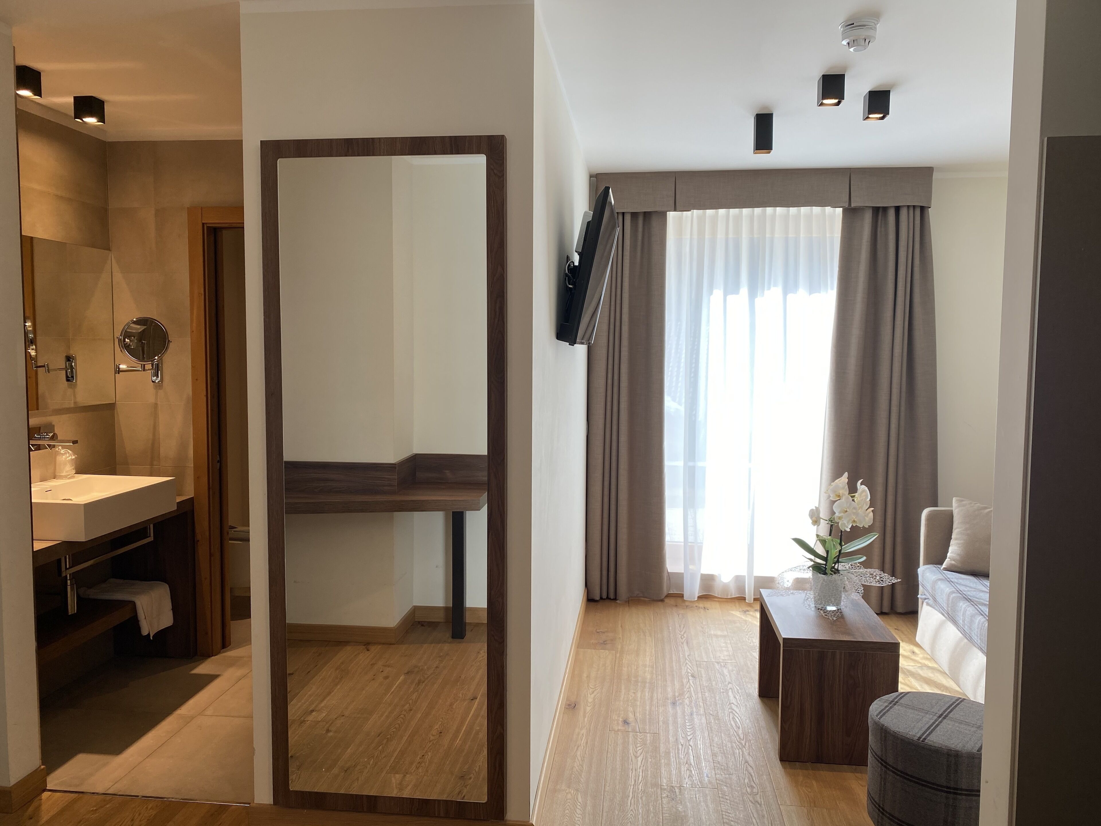 A modern hotel room with a wooden floor, a large mirror, a bathroom with a sink and mirror, a sofa, a coffee table, and a window with curtains.