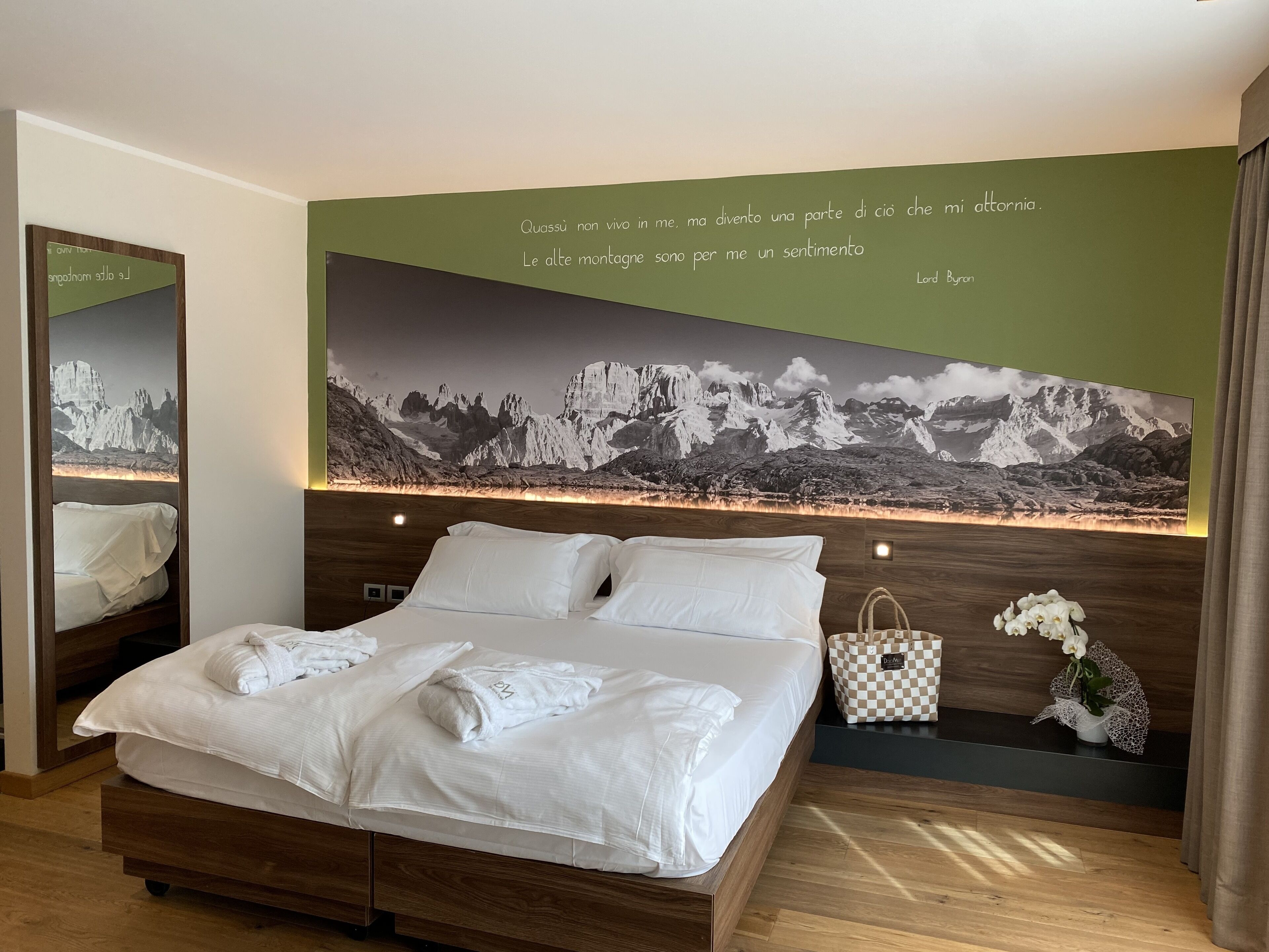 A modern hotel room with a large bed, a mirror, a wall-mounted artwork of a mountain landscape, and a small plant on the nightstand.