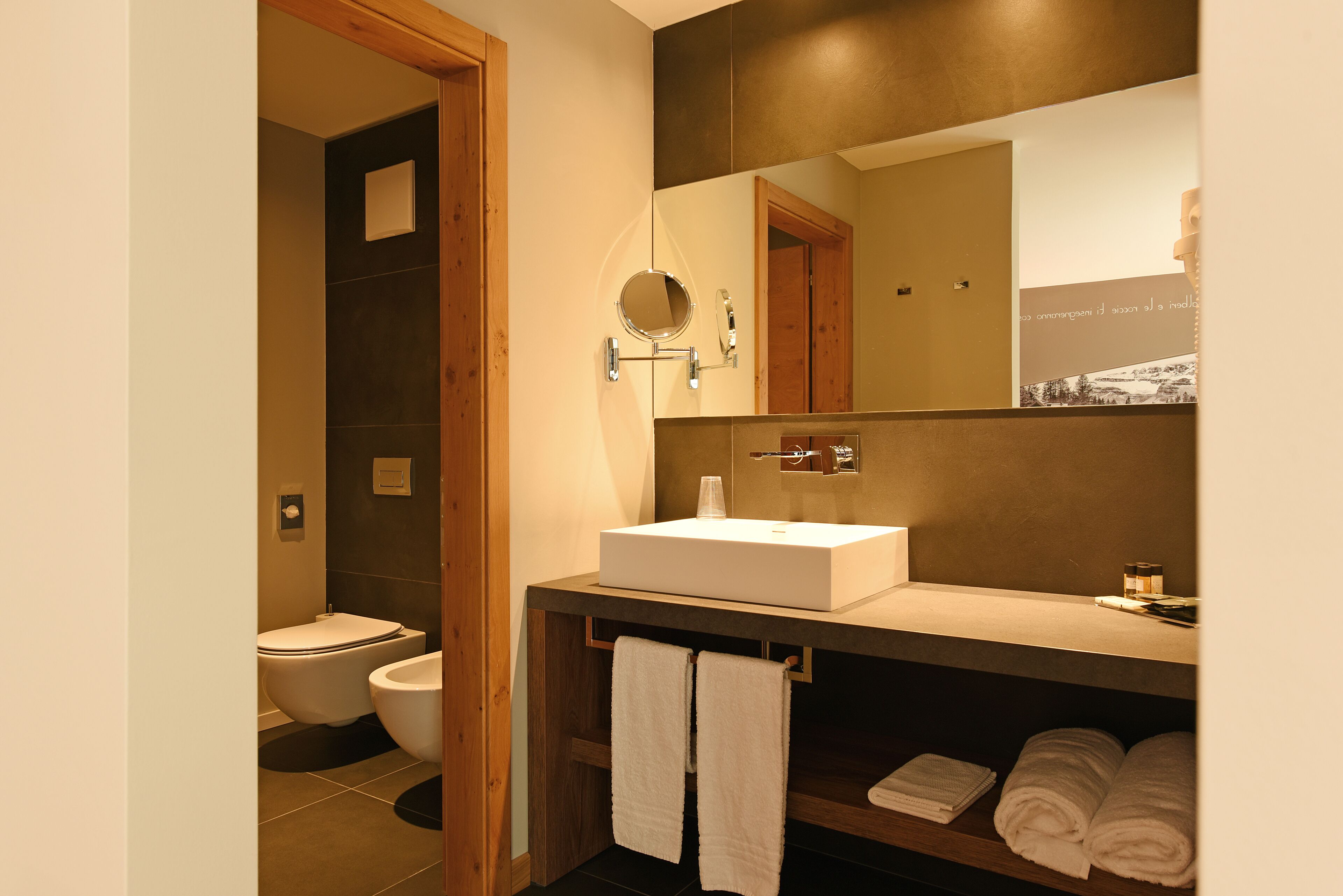 A modern bathroom with a large mirror, a white sink, and a toilet. There are towels on a shelf and a wooden door frame.