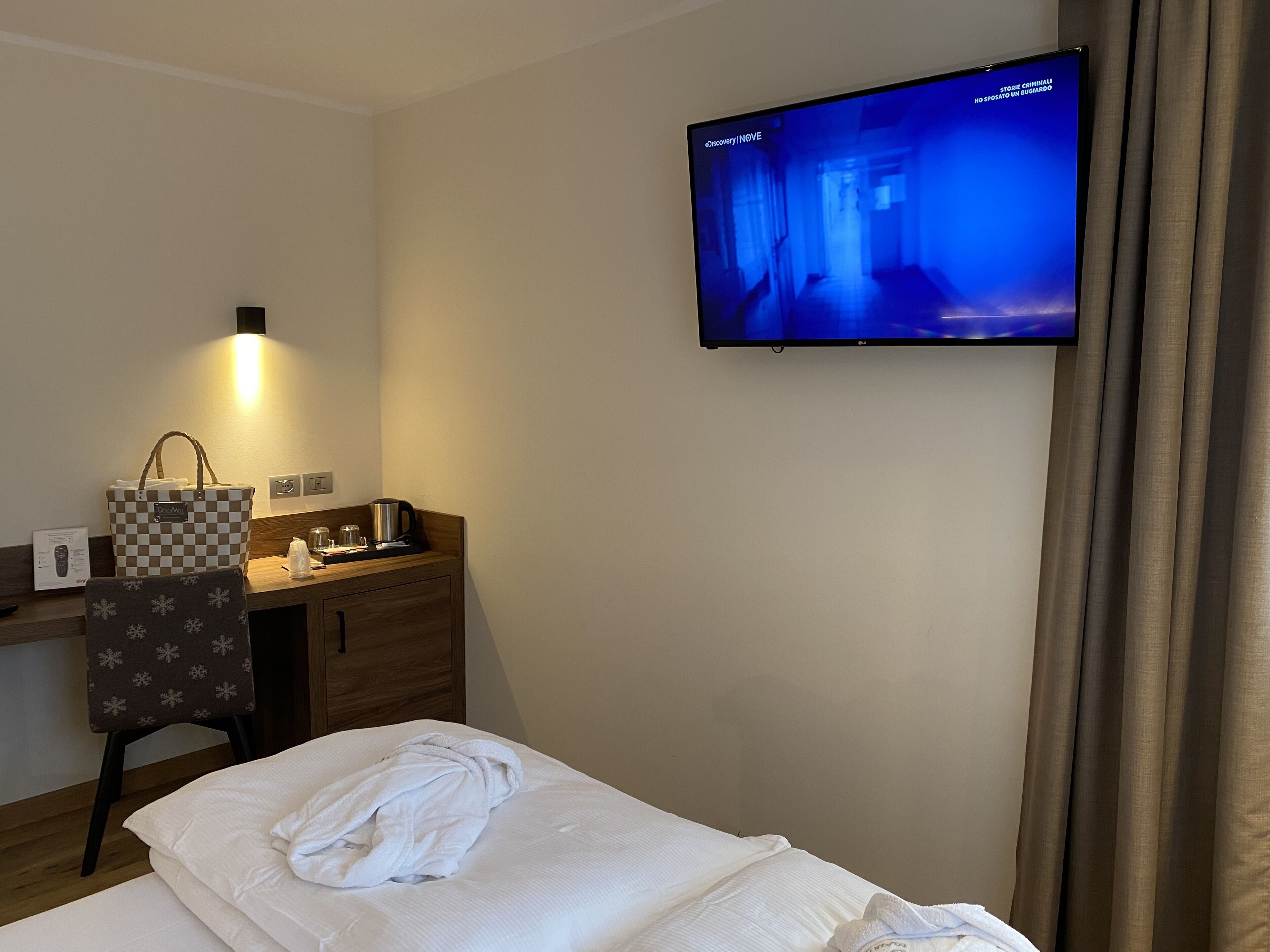 A hotel room with a bed, a desk with a computer, a television mounted on the wall, and a chair.