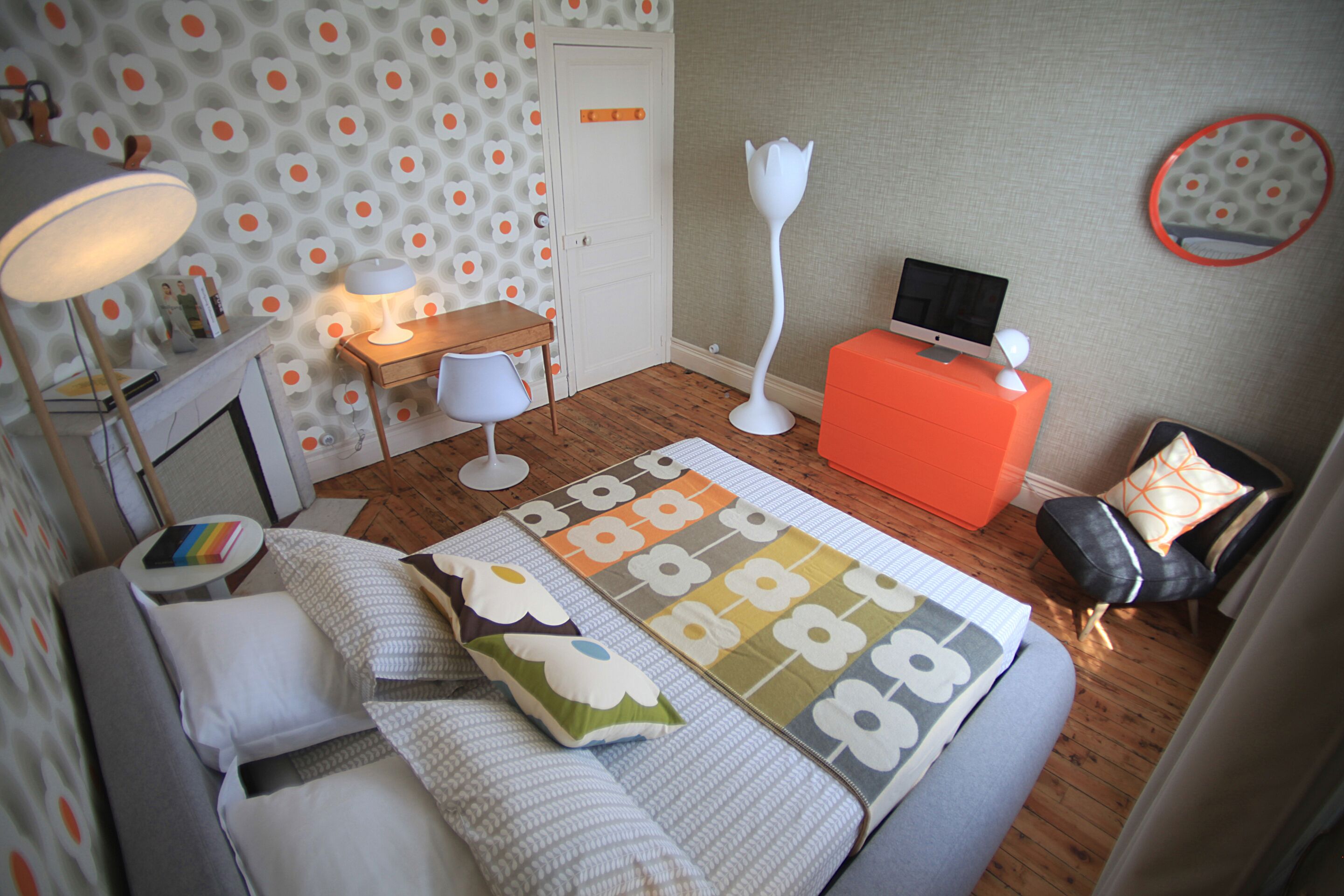 Ein Schlafzimmer mit einem Bett, einem Schreibtisch, einem Sessel, einer Stehlampe und einem Fernseher auf einem Standfuß.