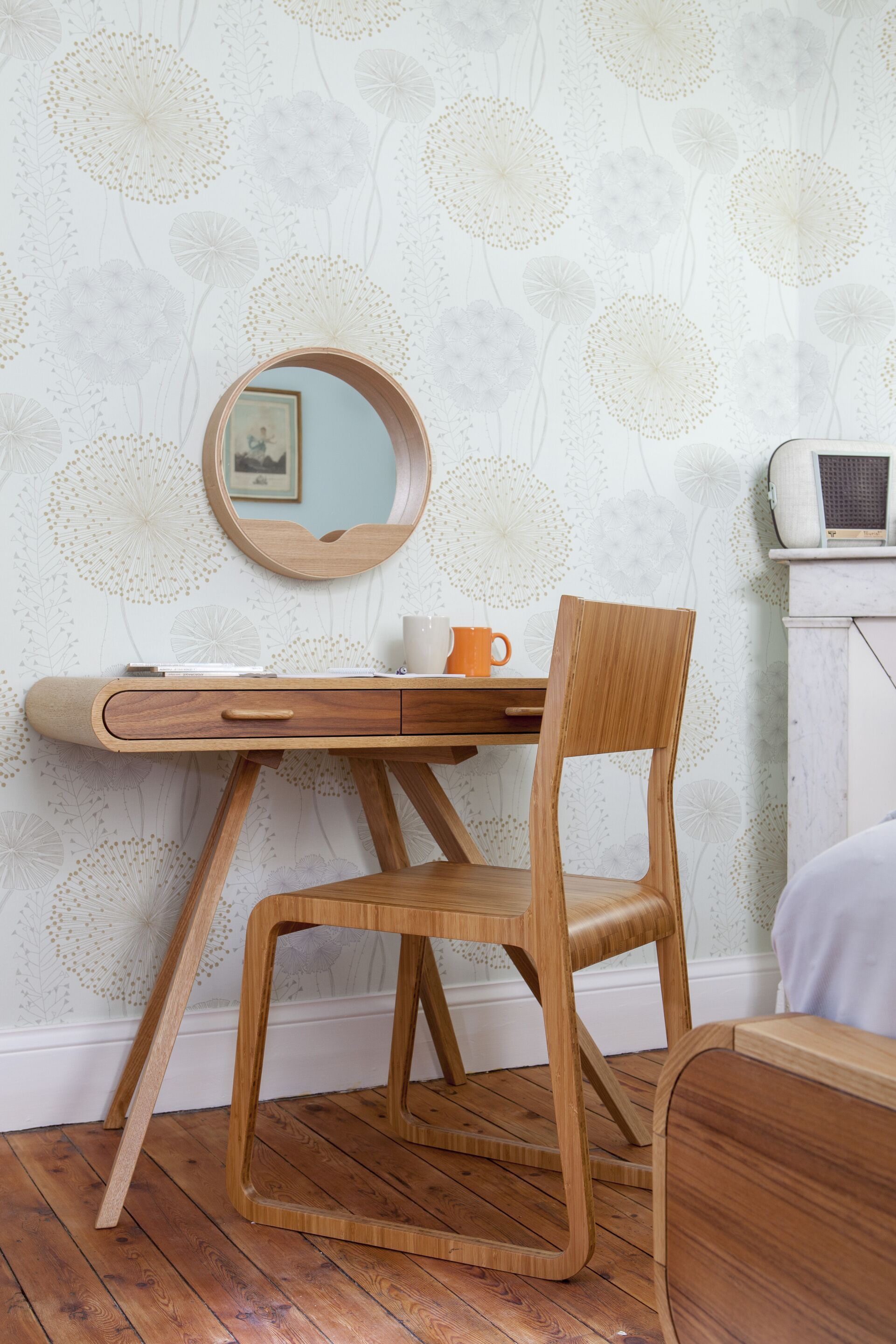 A wooden desk with a chair, a round mirror, and a small orange mug on the wall.